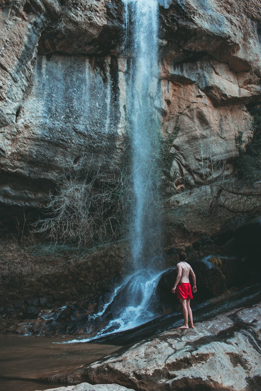 woman in red tank top and black pants standing on rock formation near waterfalls during daytime