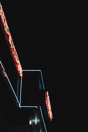 Neon lights illuminate a structure in the darkness, featuring glowing red and blue colors with visible text forming part of the signage. The background is completely black, making the neon lights stand out vividly.