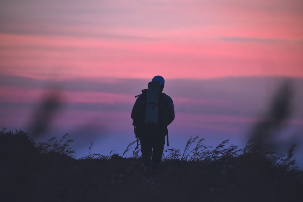 homem em jaqueta preta e mochila preta em pé no campo de grama durante o pôr do sol