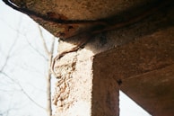 A close-up view of an aged concrete structure with visible erosion and exposed pebbles. Rusty rebar is protruding from the concrete, emphasizing the decay of the material. In the background, bare tree branches are visible against a clear sky.