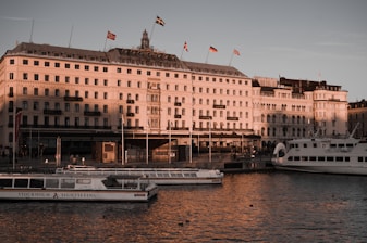 A grand building with multiple flags on the roof is bathed in warm sunlight, situated beside a calm body of water. In front of the building, several boats marked with 'Stockholm Sightseeing' float peacefully. The architecture is ornate and historic, suggesting a significant landmark or hotel.