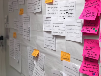 A wall is covered with various papers pinned or taped, including white sheets with handwritten notes and colored sticky notes. The notes are in different languages and appear to contain brainstorming ideas or questions. The scene conveys an organized creative space, possibly used for planning or collaboration.