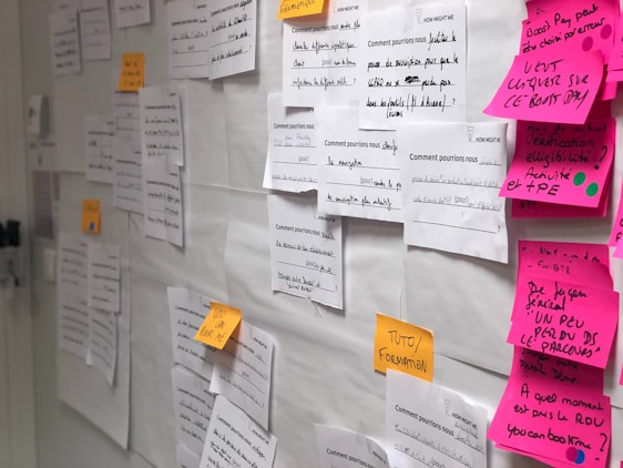 A wall is covered with various papers pinned or taped, including white sheets with handwritten notes and colored sticky notes. The notes are in different languages and appear to contain brainstorming ideas or questions. The scene conveys an organized creative space, possibly used for planning or collaboration.