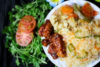 A plate of food featuring fried rice with green chili peppers, crispy glazed chicken, and mixed vegetables. Slices of tomato and fresh cilantro are arranged decoratively on the side, alongside a white flower.