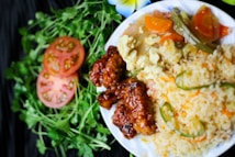 A plate of food featuring fried rice with green chili peppers, crispy glazed chicken, and mixed vegetables. Slices of tomato and fresh cilantro are arranged decoratively on the side, alongside a white flower.