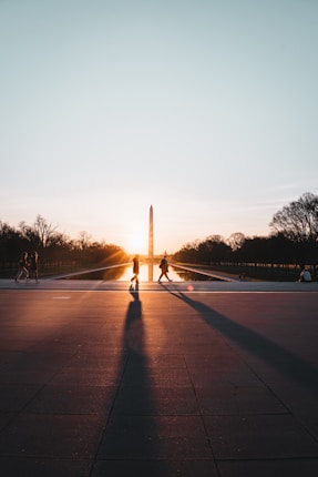 Silhouettes of people walking on a plaza during sunset with the Washington Monument in the background. The sun is low in the sky, casting long shadows on the ground, and the monument is reflected in a pool of water.