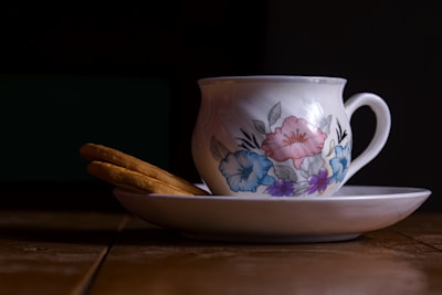 A porcelain teacup with floral designs in pink, blue, and purple rests on a matching saucer. Two biscuits are placed on the saucer beside the cup. The setting is on a wooden table with a dark background, creating a warm and cozy atmosphere.