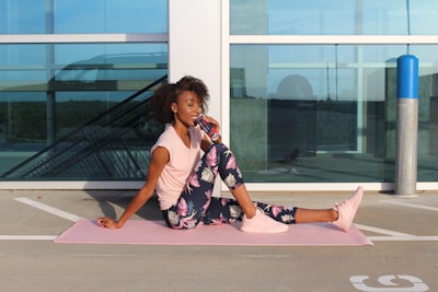 A woman is sitting on a pink yoga mat outdoors, leaning against a windowed structure. She is wearing a light pink top, floral pattern leggings, and pink sneakers while drinking from a can. The scene appears to be on a rooftop or parking area with modern architecture in the background.