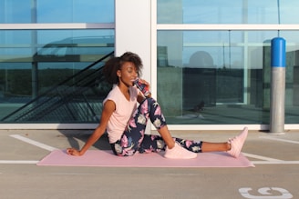 A woman is sitting on a pink yoga mat outdoors, leaning against a windowed structure. She is wearing a light pink top, floral pattern leggings, and pink sneakers while drinking from a can. The scene appears to be on a rooftop or parking area with modern architecture in the background.