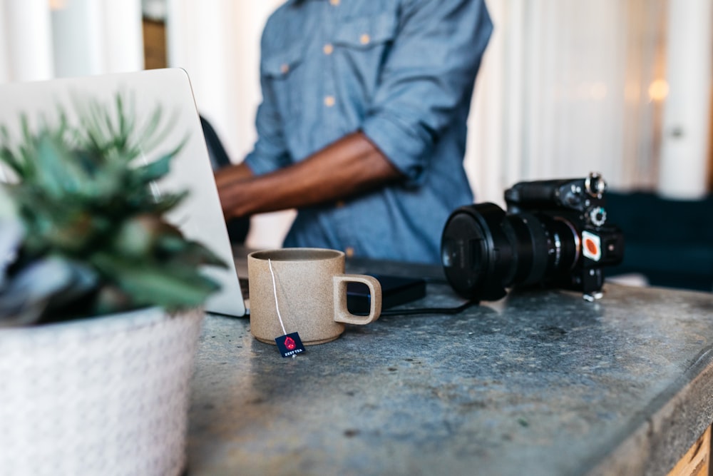 man in blue dress shirt sitting beside table with dslr camera and mug