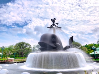 A grand fountain featuring a large sculpture of a friendly whale with a smaller figure standing atop it, set against a bright, partly cloudy sky. The fountain's water cascades gracefully in multiple tiers, creating a lively and dynamic scene surrounded by lush greenery and decorative structures.