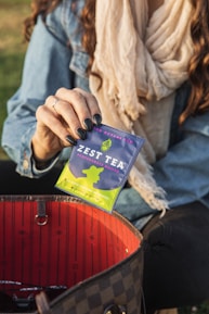 A person holding a packet of Zest Tea Pomegranate Mojito flavor with dark-painted nails. The packet is positioned above an open handbag with a red interior. The person is wearing a light scarf and denim jacket, sitting outdoors.