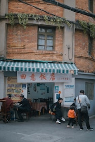 A small, casual street-side restaurant with a red brick exterior and a striped green and white awning. Several people are seated at tables outside, enjoying their meals. The wall near the entrance is decorated with menus and posters. There are plants hanging from a ledge above the entrance.