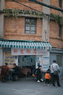 A small, casual street-side restaurant with a red brick exterior and a striped green and white awning. Several people are seated at tables outside, enjoying their meals. The wall near the entrance is decorated with menus and posters. There are plants hanging from a ledge above the entrance.