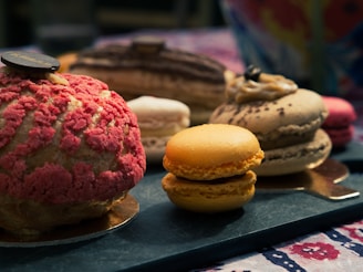 An assortment of colorful and intricately designed pastries and macarons displayed on a textured surface. The pastries exhibit a variety of textures, including a crumbly pink topping and smooth cream layers. The setting suggests a cozy and inviting atmosphere.