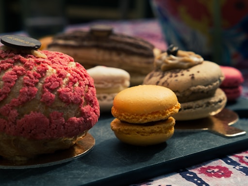 An assortment of colorful and intricately designed pastries and macarons displayed on a textured surface. The pastries exhibit a variety of textures, including a crumbly pink topping and smooth cream layers. The setting suggests a cozy and inviting atmosphere.
