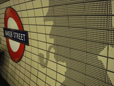 A tiled wall features a silhouette made up of smaller black tile patterns, resembling a detective smoking a pipe. To the left, a circular London Underground sign with 'Baker Street' written on a blue background stands out.