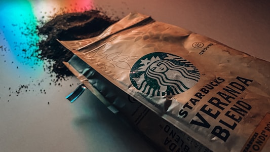 A package of Starbucks Veranda Blend coffee lies on a surface with some coffee grounds scattered nearby. The lighting casts colorful reflections on the surface, adding a dramatic effect.