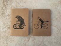 Two notebooks with kraft paper covers are placed side by side on a stone-textured surface. The notebook on the left features an illustration of a raccoon riding a bicycle, while the one on the right shows a fox riding a bicycle with a bird on its back.