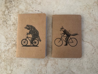 Two notebooks with kraft paper covers are placed side by side on a stone-textured surface. The notebook on the left features an illustration of a raccoon riding a bicycle, while the one on the right shows a fox riding a bicycle with a bird on its back.