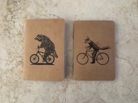 Two notebooks with kraft paper covers are placed side by side on a stone-textured surface. The notebook on the left features an illustration of a raccoon riding a bicycle, while the one on the right shows a fox riding a bicycle with a bird on its back.