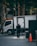 A man in a dark suit stands on a tiled urban sidewalk near a parked white delivery truck. Sunlight filters through the trees lining the street, creating patches of light and shadow. The scene also includes an orange traffic cone and some commercial signage in the background.