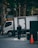 A man in a dark suit stands on a tiled urban sidewalk near a parked white delivery truck. Sunlight filters through the trees lining the street, creating patches of light and shadow. The scene also includes an orange traffic cone and some commercial signage in the background.