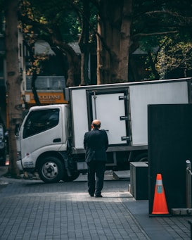 A man in a dark suit stands on a tiled urban sidewalk near a parked white delivery truck. Sunlight filters through the trees lining the street, creating patches of light and shadow. The scene also includes an orange traffic cone and some commercial signage in the background.