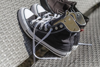 A pair of black and white high-top sneakers with laces undone, resting on a woven, textured surface, which could be a bench or chair. The sneakers have metal eyelets and a clean look.