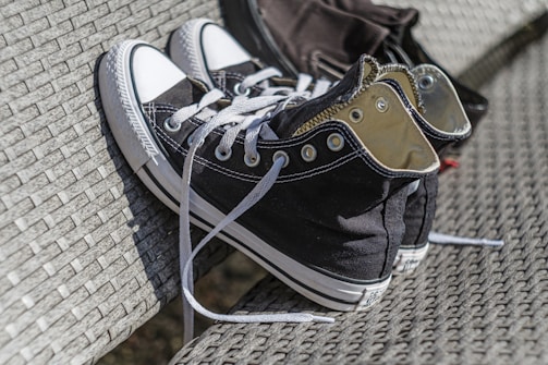 A pair of black and white high-top sneakers with laces undone, resting on a woven, textured surface, which could be a bench or chair. The sneakers have metal eyelets and a clean look.