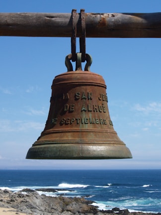 A large, weathered bronze bell hangs from a wooden beam against a backdrop of the ocean and blue sky. The bell includes lettering relating to its origin or dedication. The sea below is calm, with gentle waves crashing against the rocky shoreline.
