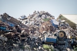 A large pile of assorted scrap metal and debris, including old vehicles, machinery parts, twisted metal sheets, and various other discarded items. The heap is situated next to a corrugated metal roof structure.