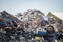 A large pile of assorted scrap metal and debris, including old vehicles, machinery parts, twisted metal sheets, and various other discarded items. The heap is situated next to a corrugated metal roof structure.