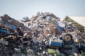 A large pile of assorted scrap metal and debris, including old vehicles, machinery parts, twisted metal sheets, and various other discarded items. The heap is situated next to a corrugated metal roof structure.