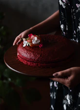 A close-up of a hand gently placing a velvety red beret-shaped cake on a rustic wooden table.