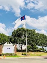 A flagpole with a flag displaying a blue field and a red cross, set against a backdrop of a white building with a tower. Surrounding the structure are lush green trees, and the scene is well-lit under a partly cloudy blue sky. A sign with the word 'WELCOME' is visible near two orange traffic cones.
