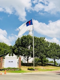 A flagpole with a flag displaying a blue field and a red cross, set against a backdrop of a white building with a tower. Surrounding the structure are lush green trees, and the scene is well-lit under a partly cloudy blue sky. A sign with the word 'WELCOME' is visible near two orange traffic cones.