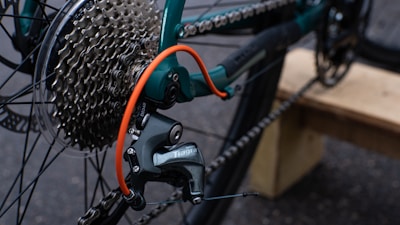 The image features a close-up view of a bicycle's gear and chain system. The gear cassette is prominently visible along with the derailleur marked with a brand name. A bright orange cable stands out against the metal components. The background is blurry, with hints of a wooden surface and asphalt.