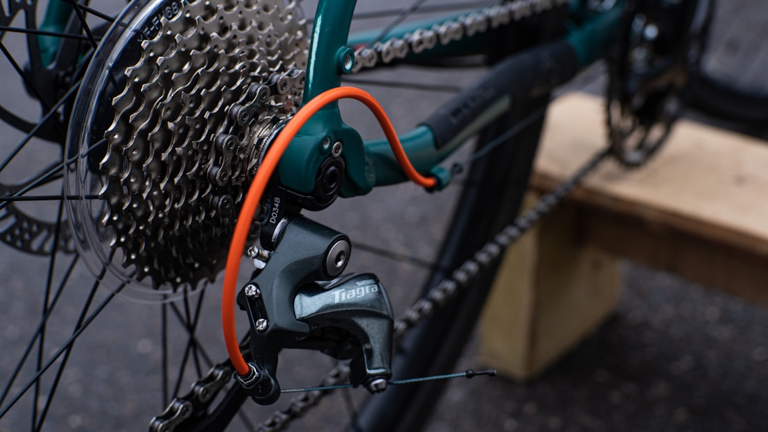 The image features a close-up view of a bicycle's gear and chain system. The gear cassette is prominently visible along with the derailleur marked with a brand name. A bright orange cable stands out against the metal components. The background is blurry, with hints of a wooden surface and asphalt.