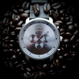 A sleek, metallic wristwatch with a contemporary design is placed amid a scattering of dark coffee beans. The watch face features three smaller dials and a red second hand, while the brand name is visible near the top. The coffee beans create a rich and textured background, accentuating the watch's shiny surface.
