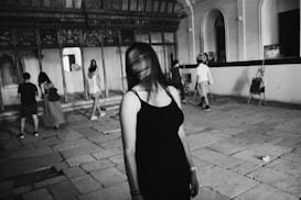 A group of people in a spacious, historic-looking room with an ornate wall and large windows. A person in the foreground has a blurred face, suggesting movement or distortion. Some individuals appear to be examining or interacting with items within the room, which include easels along the walls.