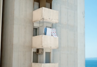 A tall concrete building features a small balcony with laundry hanging out to dry. The balcony is simple with utilitarian design, and a blue and patterned towel are visible. An air conditioning unit is positioned below the balcony window. The background shows a clear blue sky and hints of the sea, suggesting a coastal location.