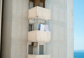 A tall concrete building features a small balcony with laundry hanging out to dry. The balcony is simple with utilitarian design, and a blue and patterned towel are visible. An air conditioning unit is positioned below the balcony window. The background shows a clear blue sky and hints of the sea, suggesting a coastal location.