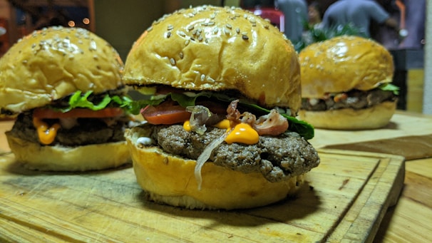 Three appetizing hamburgers are placed on a wooden board. Each burger consists of a golden, sesame-seed bun with a beef patty, fresh lettuce, slices of tomato, and melted cheese visible. The golden hue of the buns and the vibrant green of the lettuce create an appealing contrast. In the background, there's a hint of a cozy indoor setting.