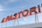 A red neon sign with bold, block letters spelling out part of a word. The sign is mounted on a white structure and is set against a backdrop of a clear blue sky. The letters are connected with yellow metal supports.