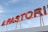 A red neon sign with bold, block letters spelling out part of a word. The sign is mounted on a white structure and is set against a backdrop of a clear blue sky. The letters are connected with yellow metal supports.