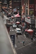 A bustling urban street scene features several people walking along the sidewalk. Red lanterns hang above the street, adding a traditional aesthetic. There are multiple shops and restaurants with signs in diverse languages, and vehicles, including a van, navigate the road. The environment is busy and multicultural.