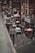 A bustling urban street scene features several people walking along the sidewalk. Red lanterns hang above the street, adding a traditional aesthetic. There are multiple shops and restaurants with signs in diverse languages, and vehicles, including a van, navigate the road. The environment is busy and multicultural.