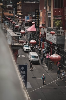 A bustling urban street scene features several people walking along the sidewalk. Red lanterns hang above the street, adding a traditional aesthetic. There are multiple shops and restaurants with signs in diverse languages, and vehicles, including a van, navigate the road. The environment is busy and multicultural.