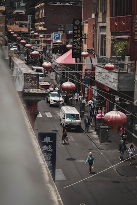 A bustling urban street scene features several people walking along the sidewalk. Red lanterns hang above the street, adding a traditional aesthetic. There are multiple shops and restaurants with signs in diverse languages, and vehicles, including a van, navigate the road. The environment is busy and multicultural.